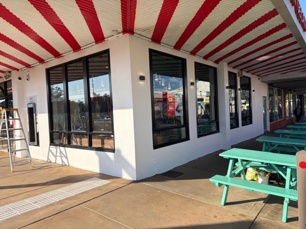Corner view of a white storefront with large windows under a red and cream striped awning, and teal picnic tables outside.