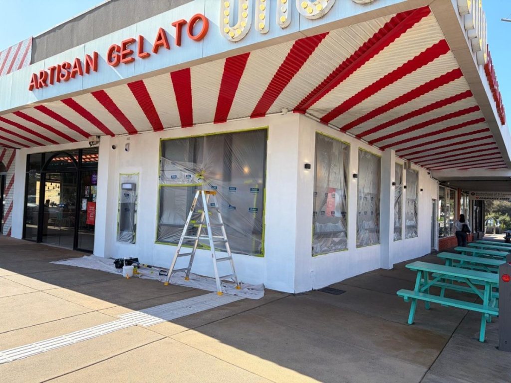 Exterior of a gelato shop under renovation; plastic-covered windows, ladder and painting supplies, red-and-white striped awning, and turquoise picnic tables.