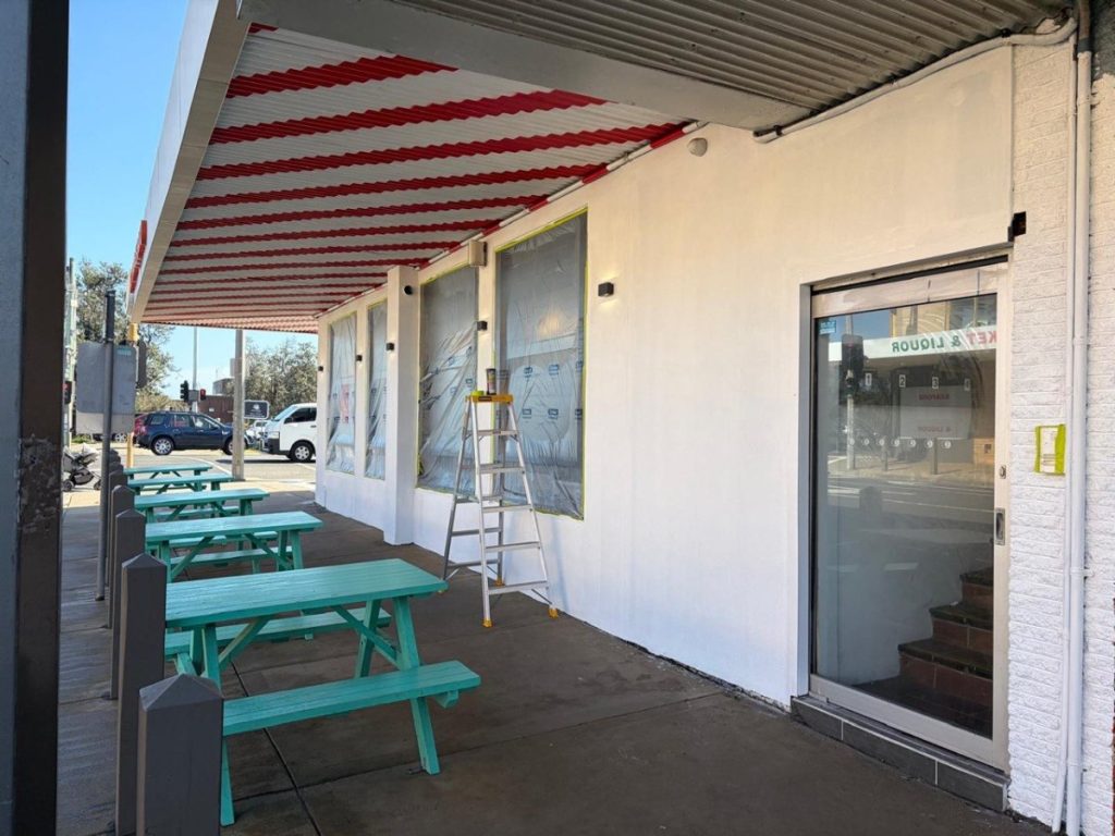 Storefront under renovation with plastic-covered windows, a ladder, and teal picnic tables outside under a red-striped awning