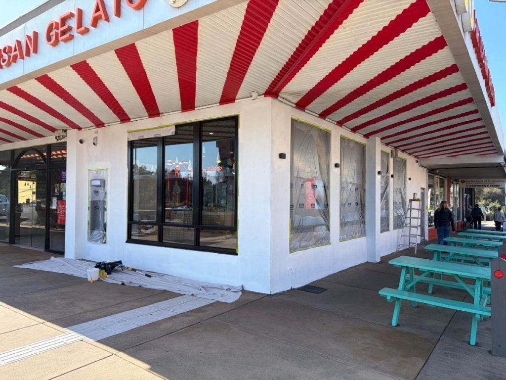 Storefront under renovation with red and white striped awning, plastic-covered windows, and teal picnic tables outside.