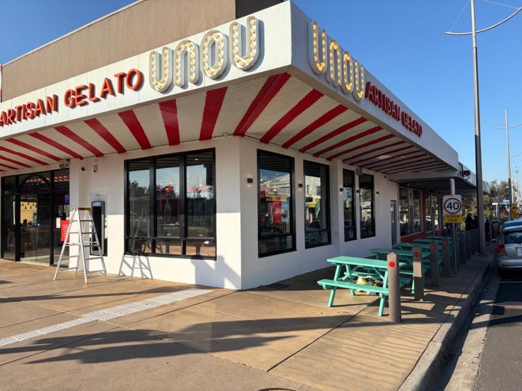 Exterior of an artisan gelato shop with a red striped awning, large glass windows, and turquoise picnic tables outside on a sunny day.