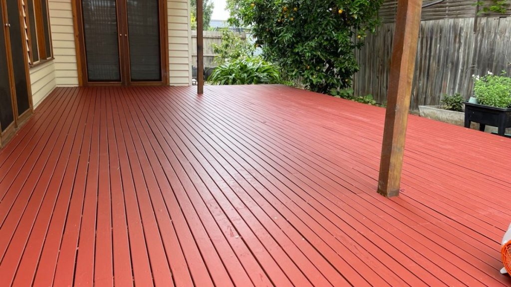 Red-painted wooden deck extending from a house with sliding doors and wooden posts, bordered by a fence and greenery in the background.