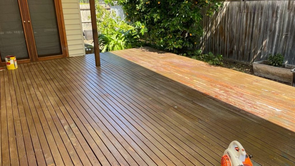 Outdoor wooden deck being stained: dark brown left side, lighter bare wood right side, sliding door on the left and plants in the background.