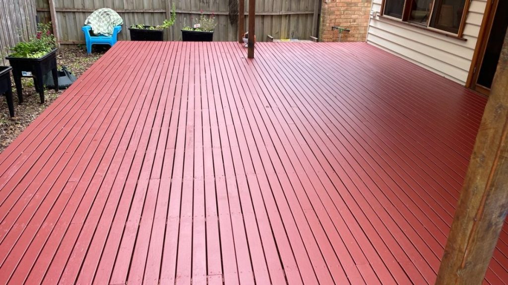 Red-painted wooden deck in a backyard, with planters along a wooden fence and a blue chair in the corner near a covered area by the house.