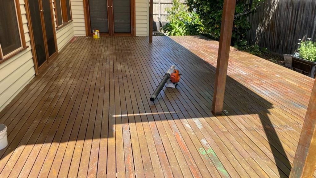 Orange leaf blower resting on a wet wooden deck beside a house, with doors and garden in the background.