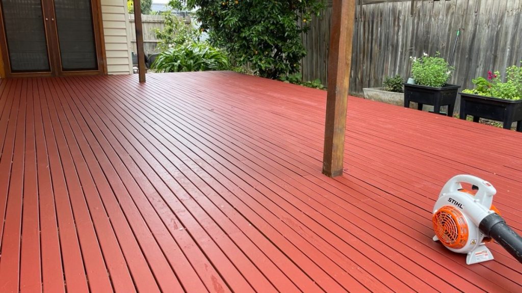 Red wooden deck underway with a STIHL leaf blower resting on the right side, plants and fence in the background.