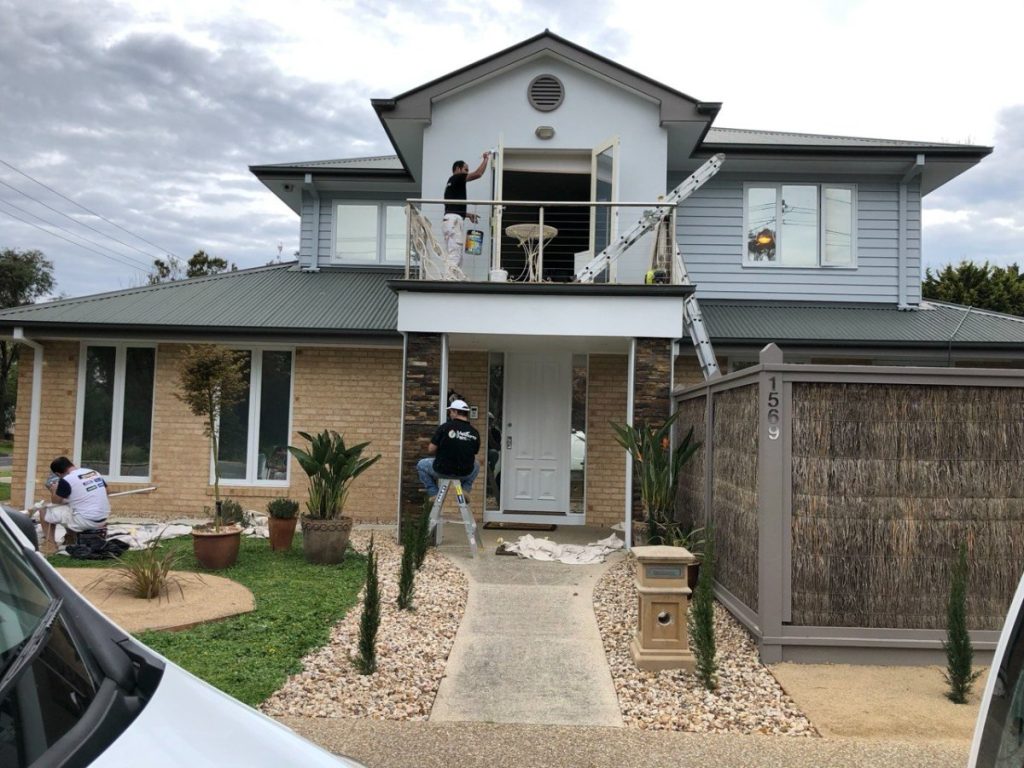 Two-story house under renovation with workers on a balcony scaffold and a ladder, front yard landscaping, and construction materials around the entrance