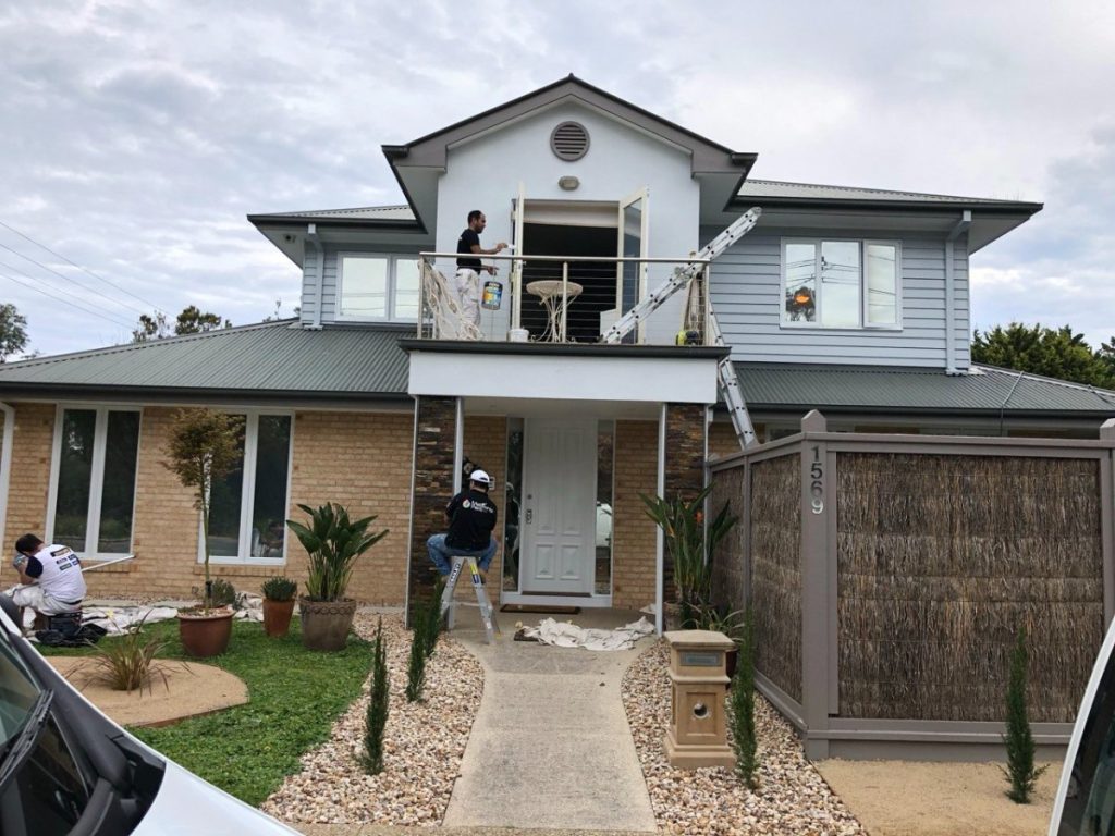 Two workers remodel a two‑story house; one on the balcony with a paint can, another on a ladder near the front door.