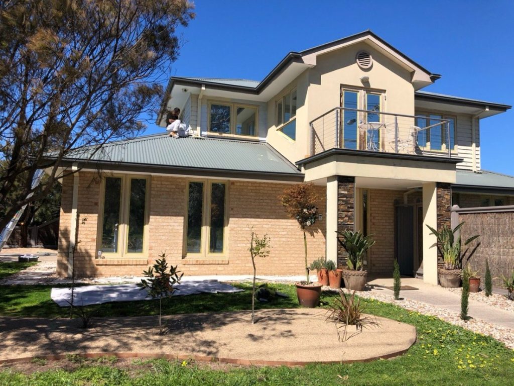 Two-story beige house with a man on the metal roof repairing it; front yard with potted plants.