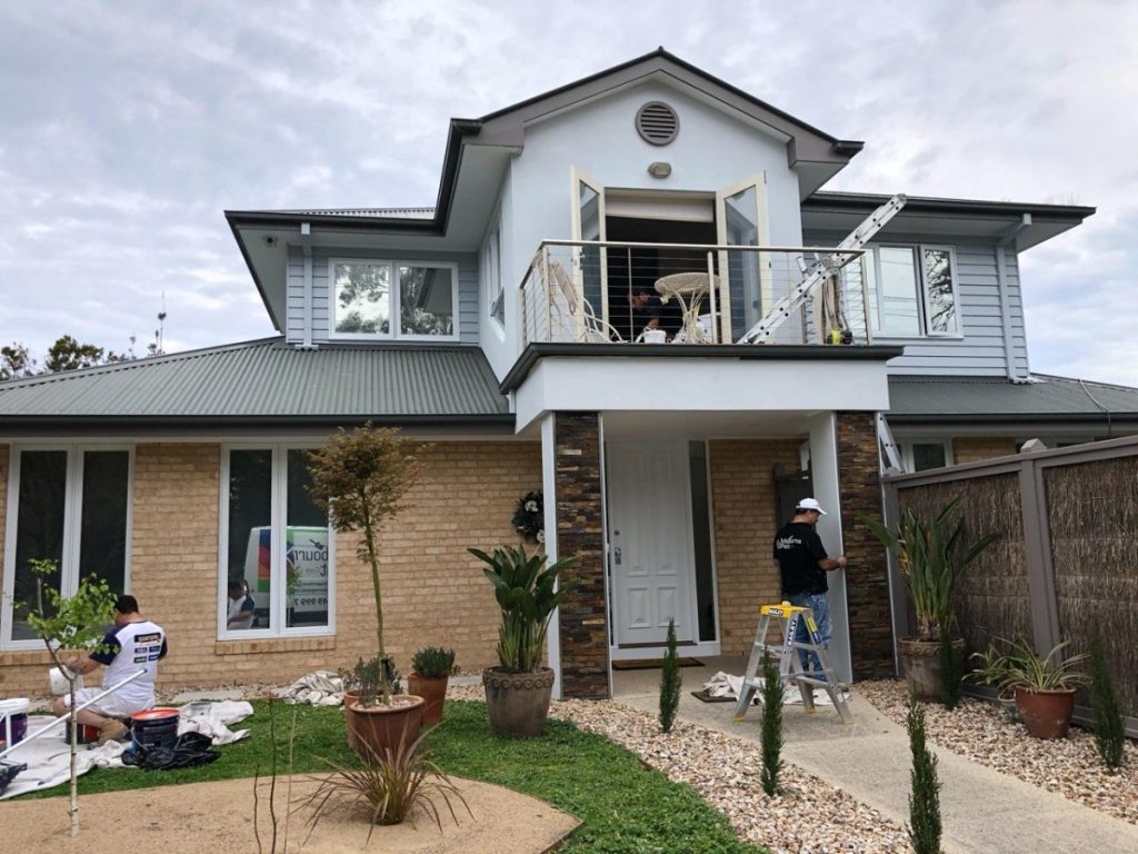 Two-story house with a balcony; workers renovating the balcony, ladder leaning on the wall, and front-yard plants nearby.