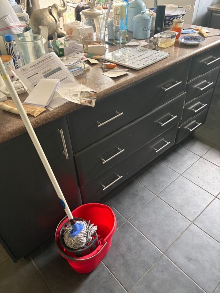 Messy kitchen counter with papers, jars, and a cluttered workspace, plus a mop and red mop bucket on the tiled floor nearby.