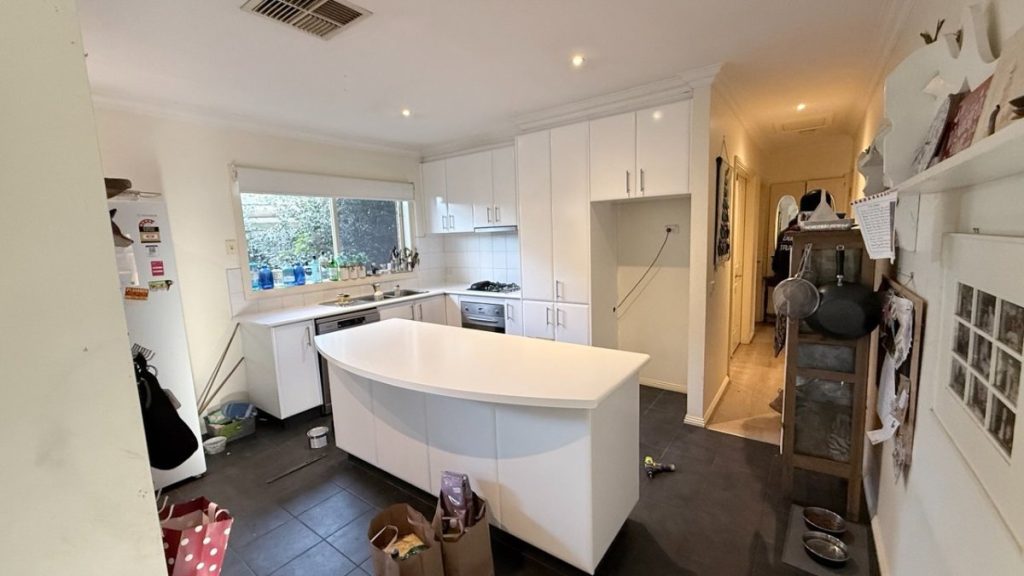 White kitchen with curved island, white cabinets, and a windowed sink area; bags and clutter on the floor nearby.