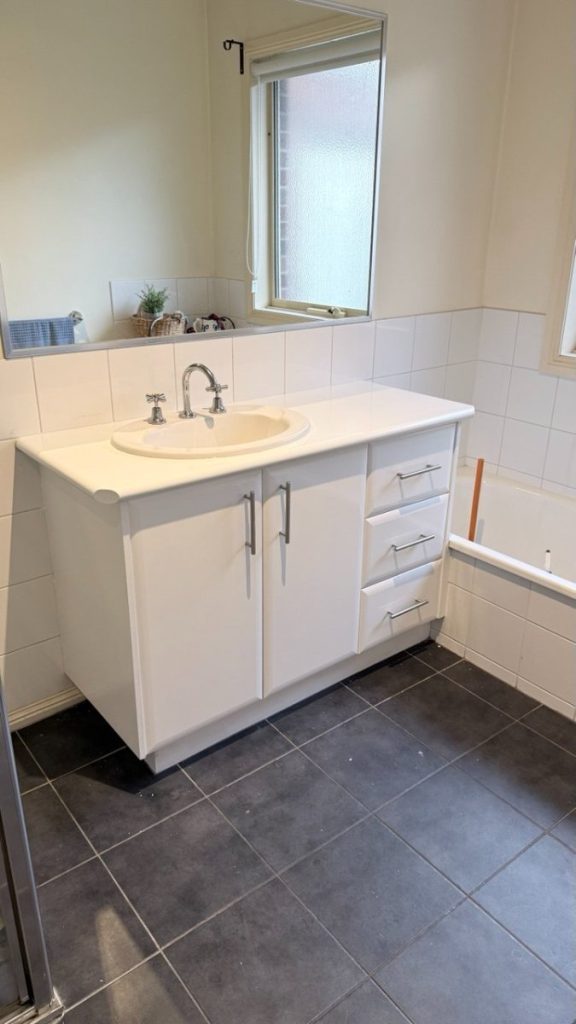 White bathroom vanity with oval sink, chrome faucet, and two-door cabinet plus three drawers under a large wall mirror; tiled backsplash and dark floor tiles visible.