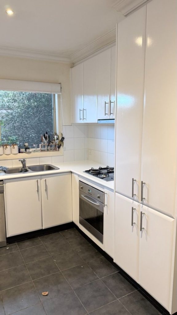 Modern white kitchen with a gas cooktop, built-in oven, and tall cabinets along a dark tile floor; sink under a window with plants on the sill.