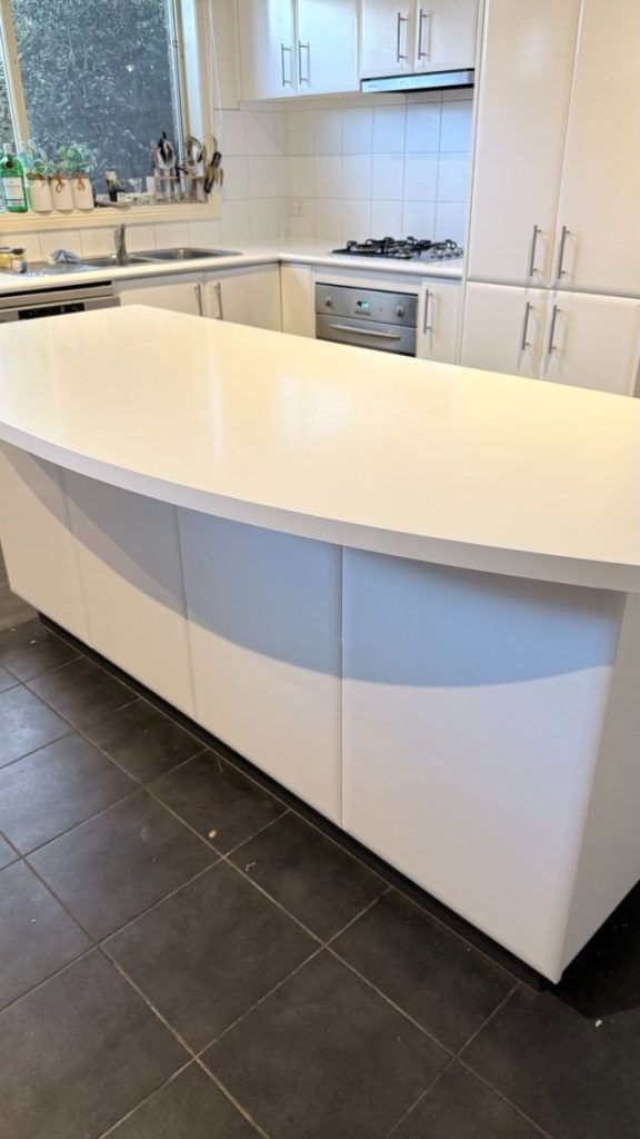 Modern white kitchen with curved island, white cabinetry, gas cooktop, built-in oven, and sink near a window.
