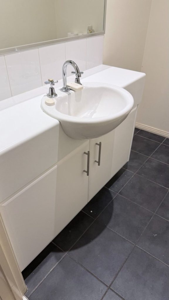 White bathroom vanity with built-in sink and chrome faucet, mirror above, dark tiled floor, and cabinets below for storage