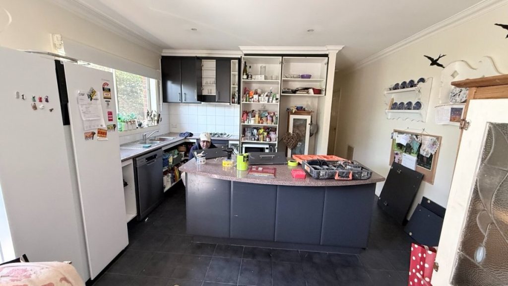 A home kitchen with a dark island, open shelves, and a person using a laptop at the counter.