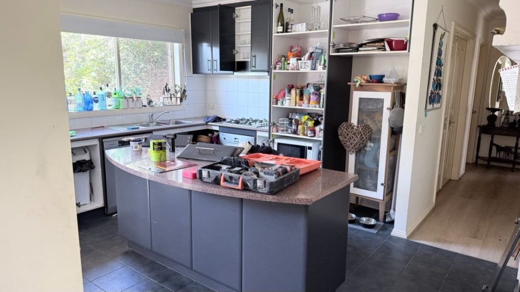 Kitchen with dark cabinets and a curved island; toolbox and tools spread across the counter, shelves stocked with supplies nearby.